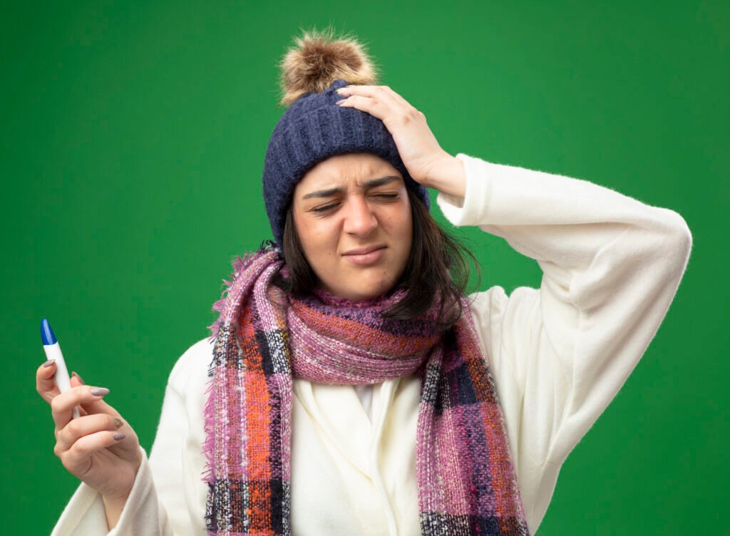 aching young caucasian ill girl wearing robe winter hat and scarf holding thermometer keeping hand on head with closed eyes isolated on green background